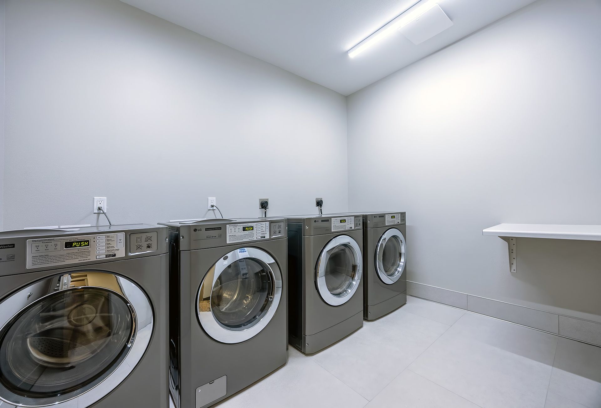 Laundry room with four washing machines and a shelf against a gray wall.