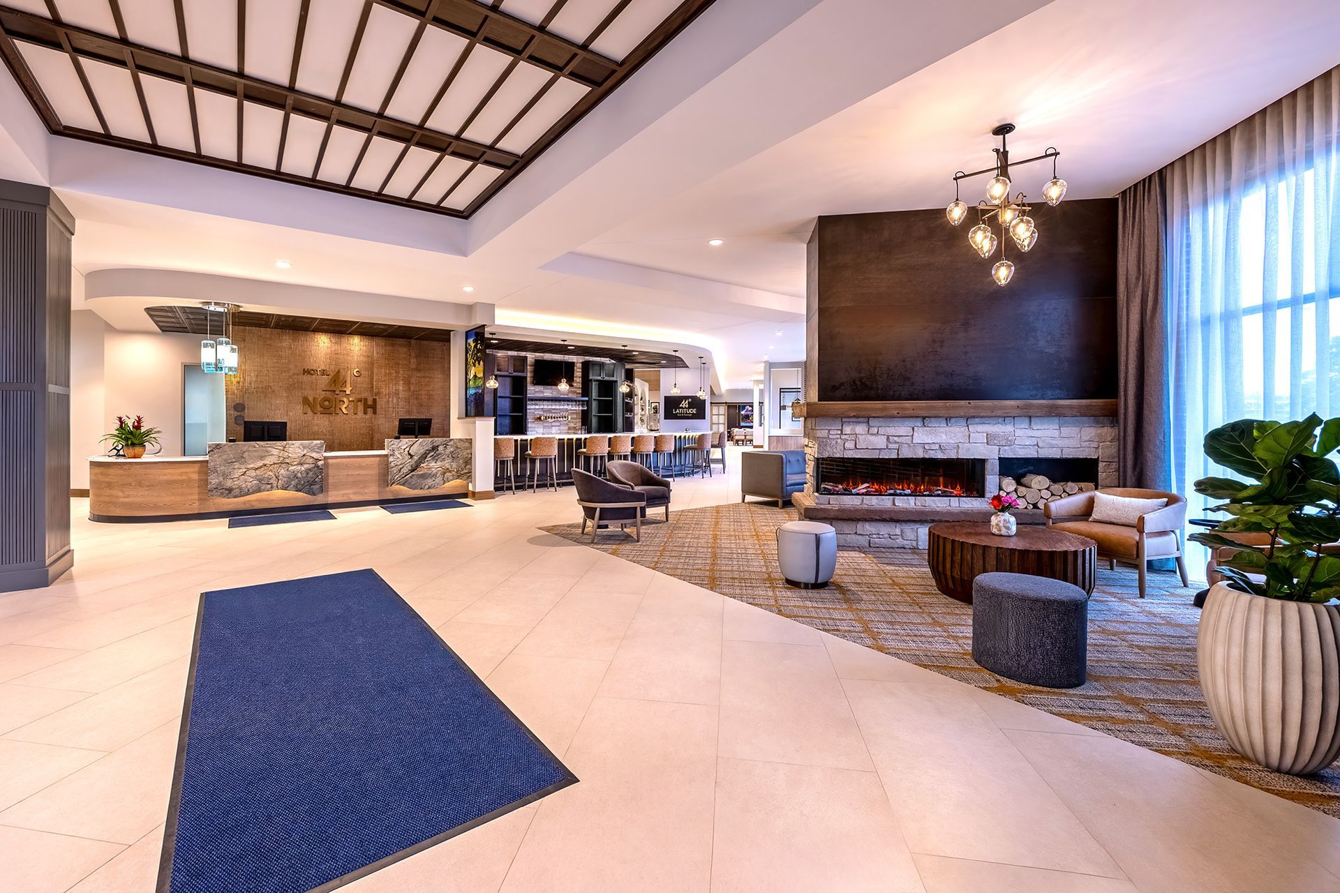 Hotel lobby with check-in desk, fireplace, seating area, and potted plant on a tiled floor.