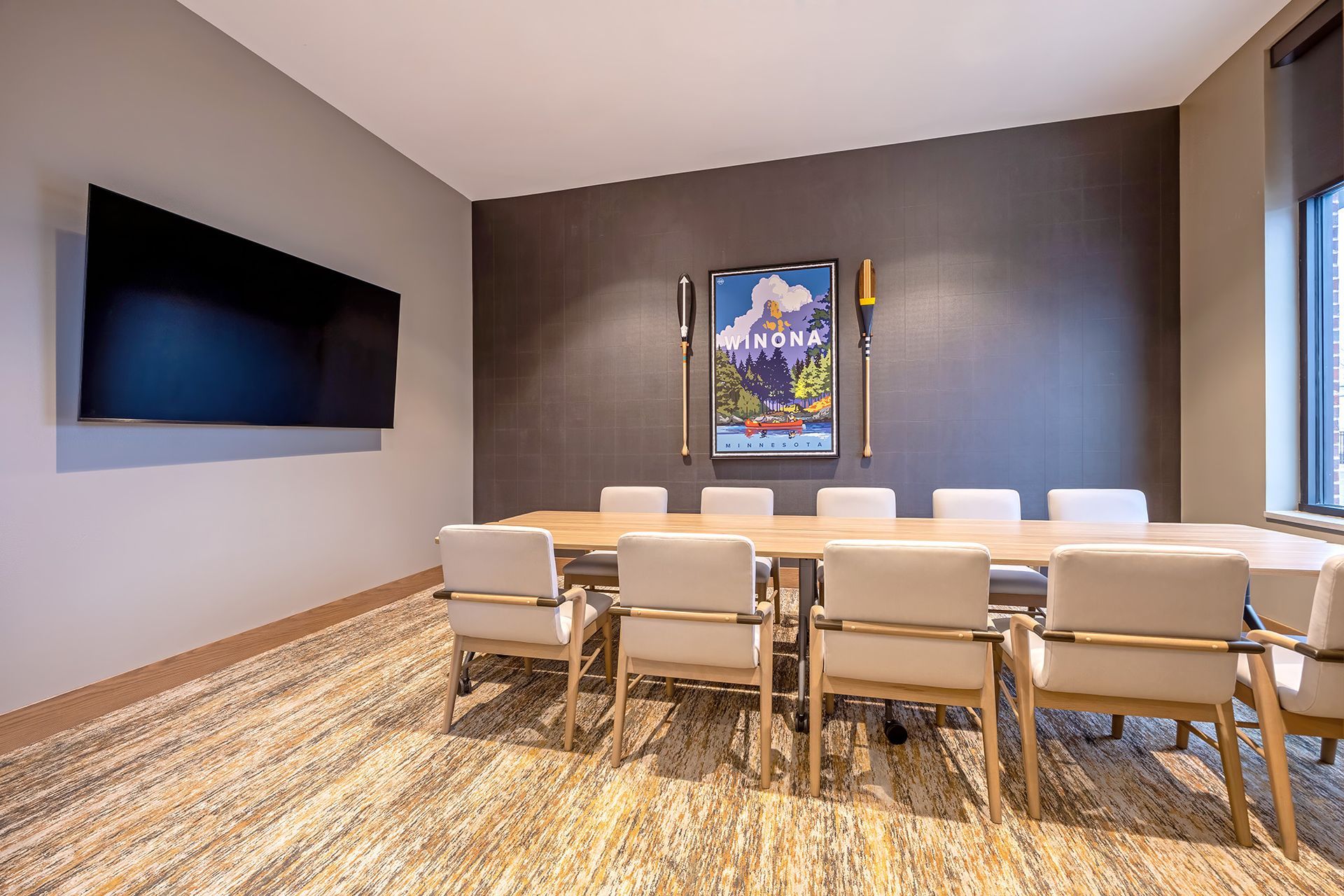 Conference room: long wood table, beige chairs, TV, dark accent wall with artwork, patterned carpet.