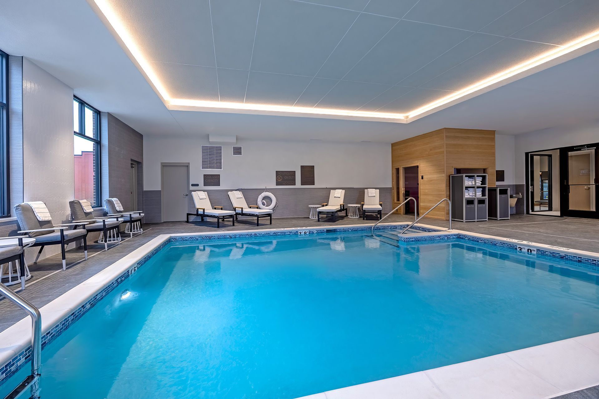 Indoor swimming pool with lounge chairs, gray walls, and bright blue water.