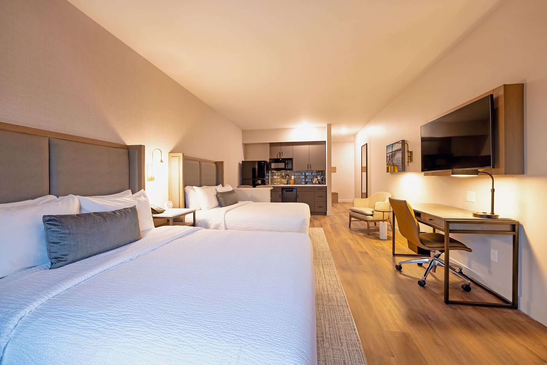 Hotel room with two beds, kitchenette, desk, and TV. Wooden floors and neutral color palette.