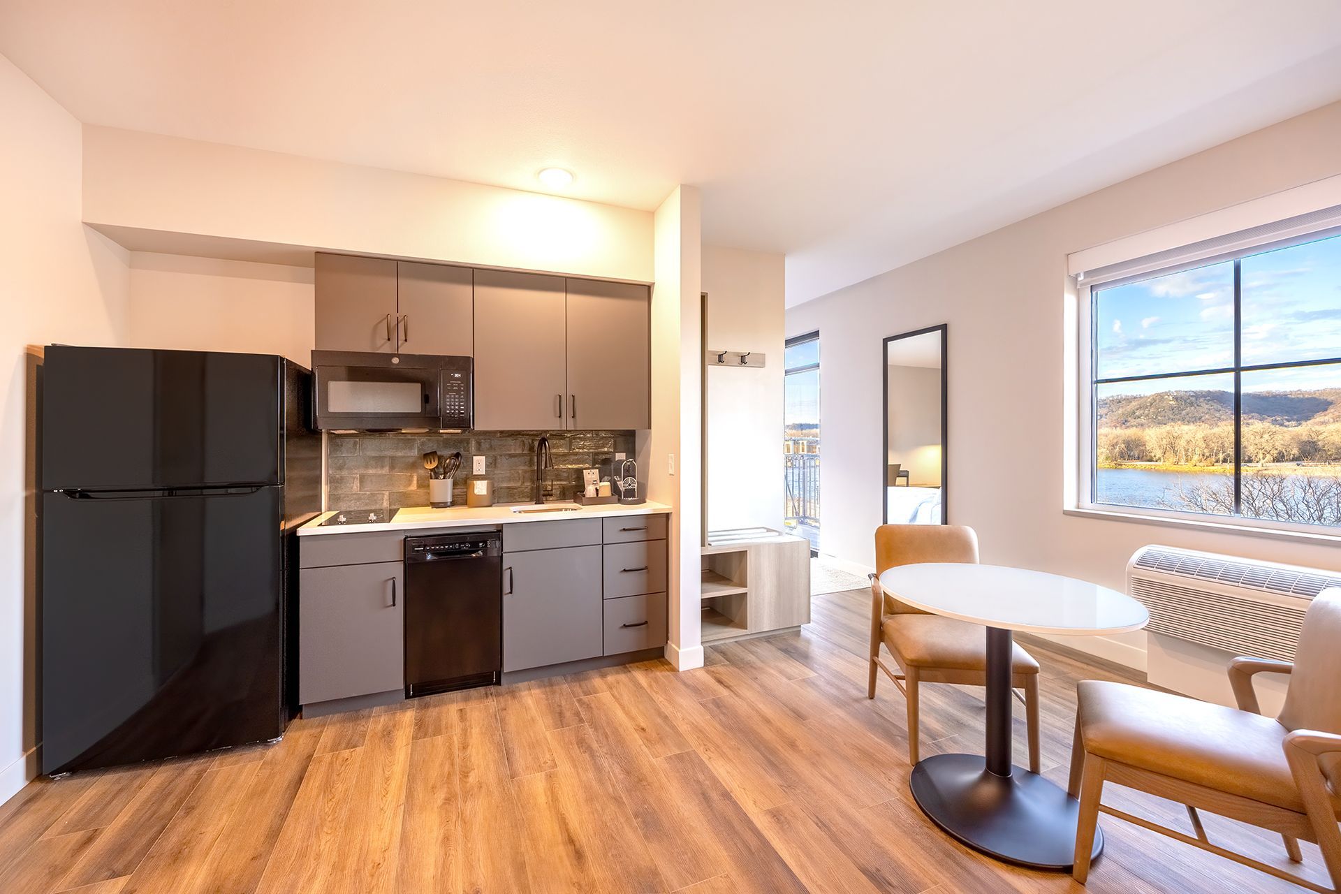 A bright kitchenette with a black refrigerator, grey cabinets, and a dining table by a window overlooking a river.