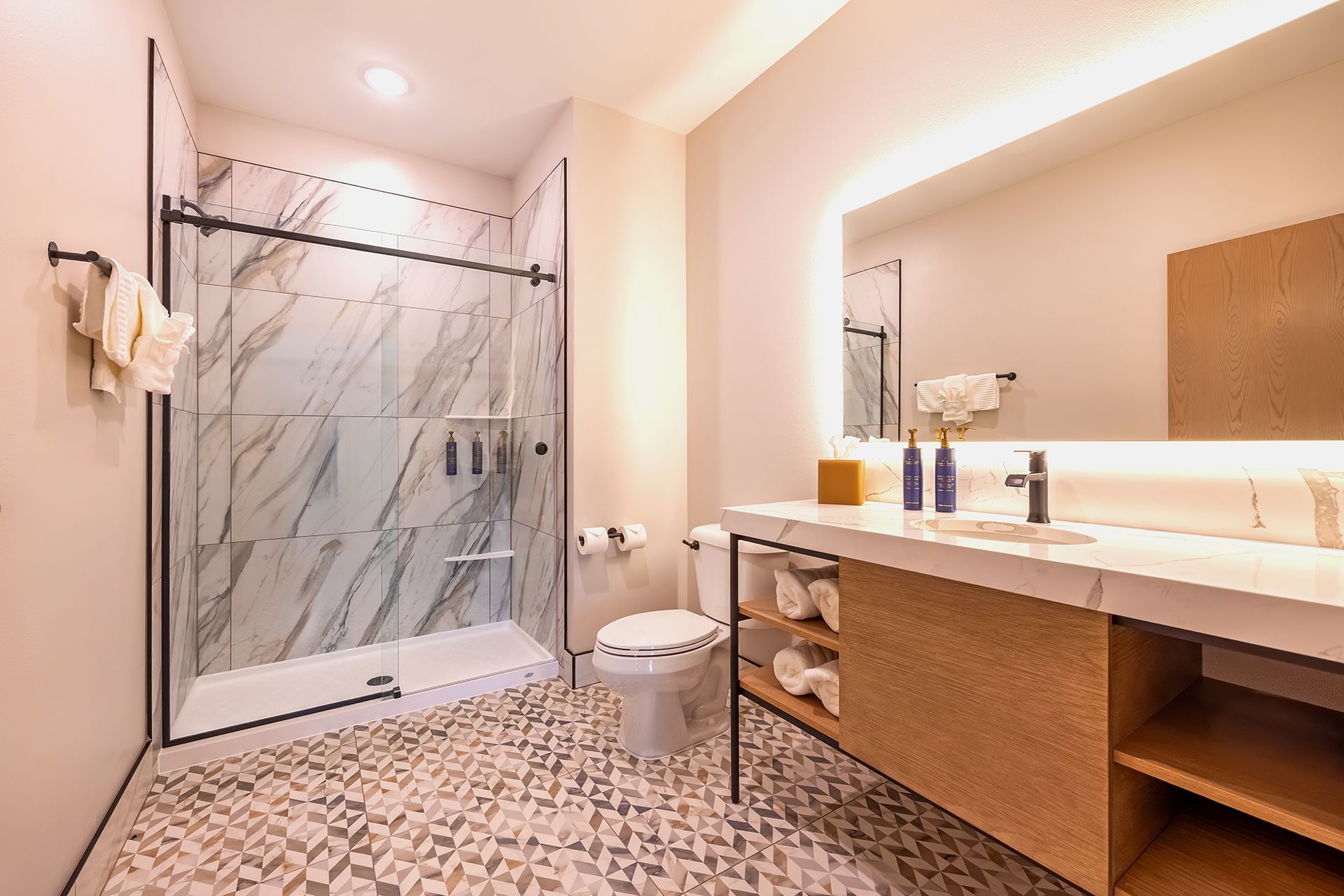 A modern bathroom features a walk-in glass shower with marble walls, a white toilet, and a vanity with a backlit mirror.