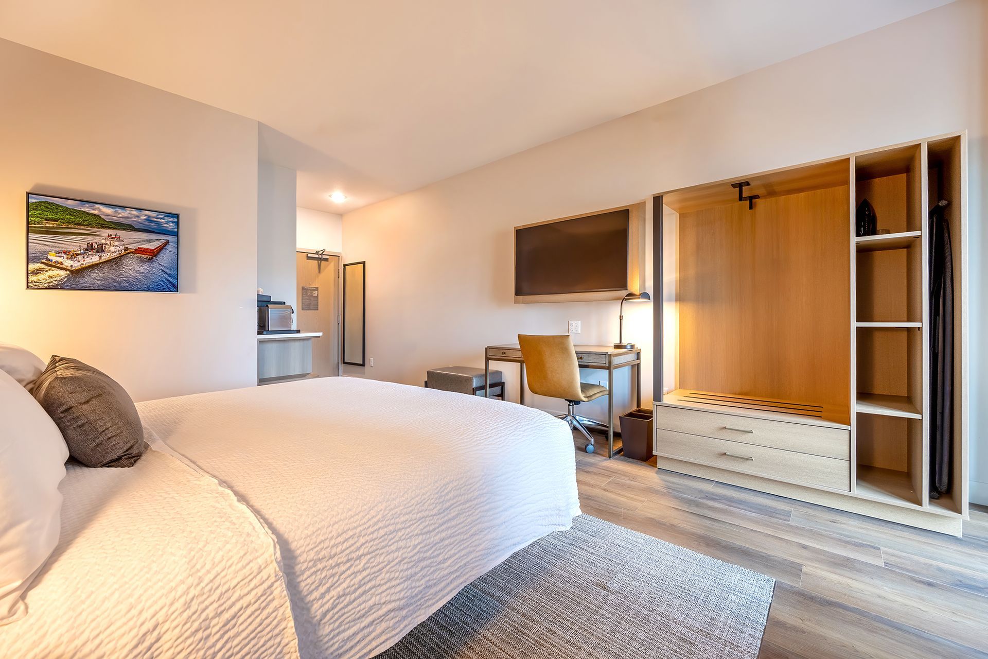 Hotel room interior: bed, desk, wardrobe, painting on wall. Neutral colors, modern design.