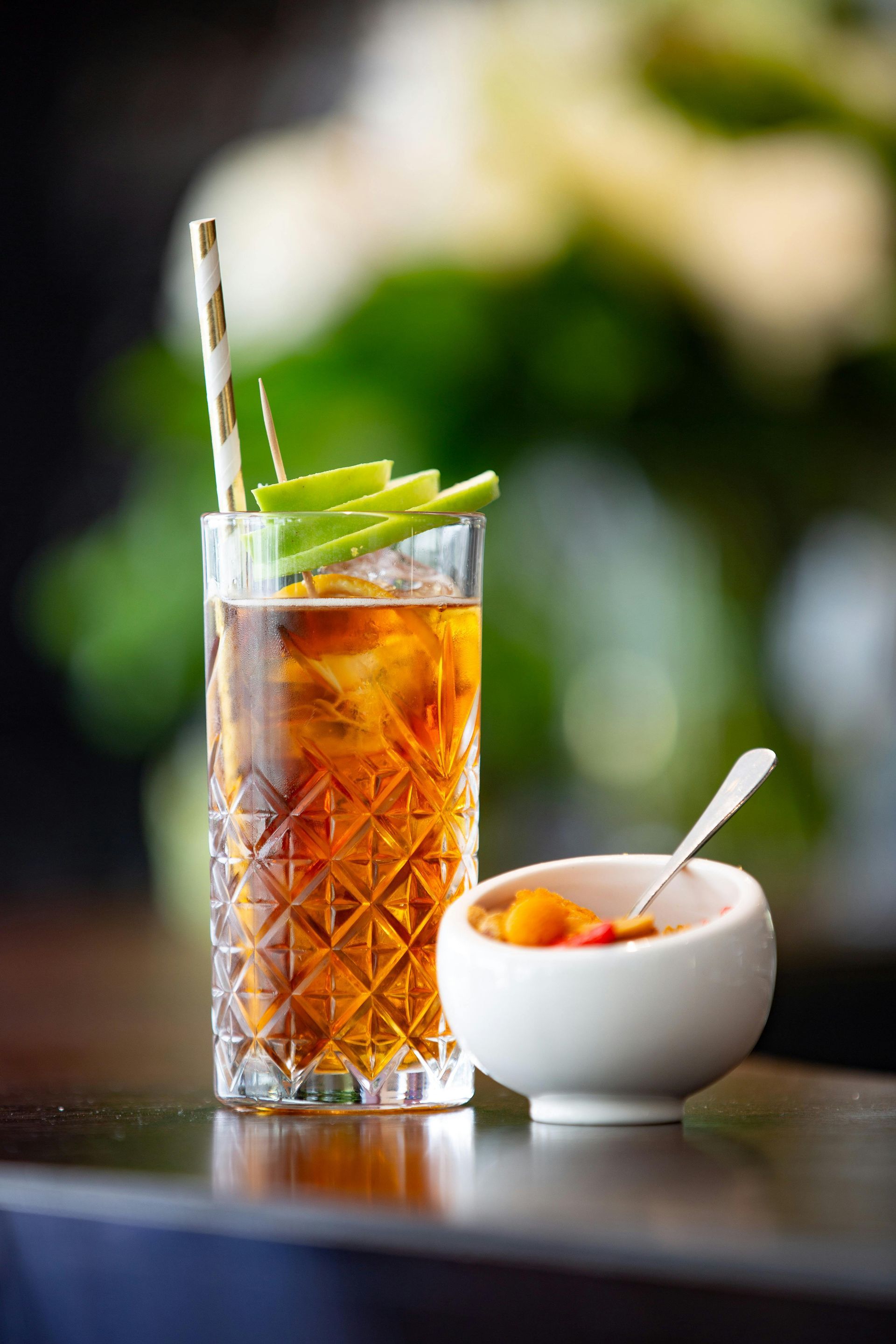 Tall glass of brown cocktail with ice, celery garnish, and small bowl of pickled vegetables.