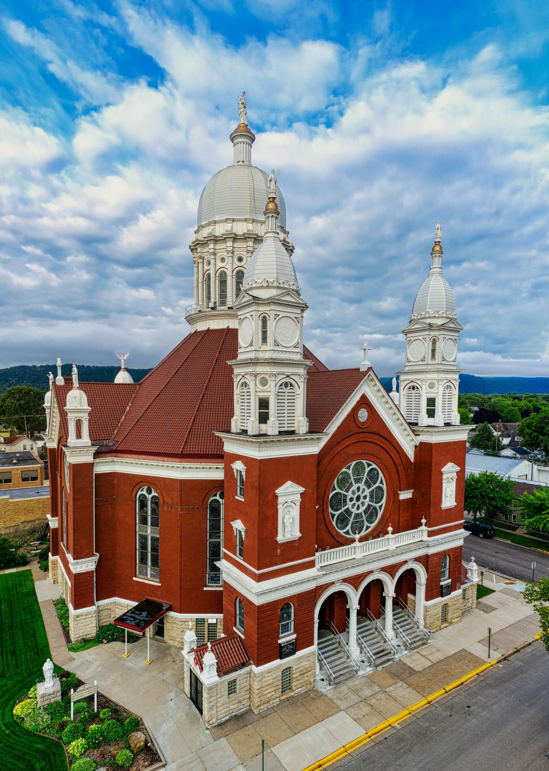 Red brick church with white trim and multiple spires under a cloudy blue sky. St. Stan's Catholic Church