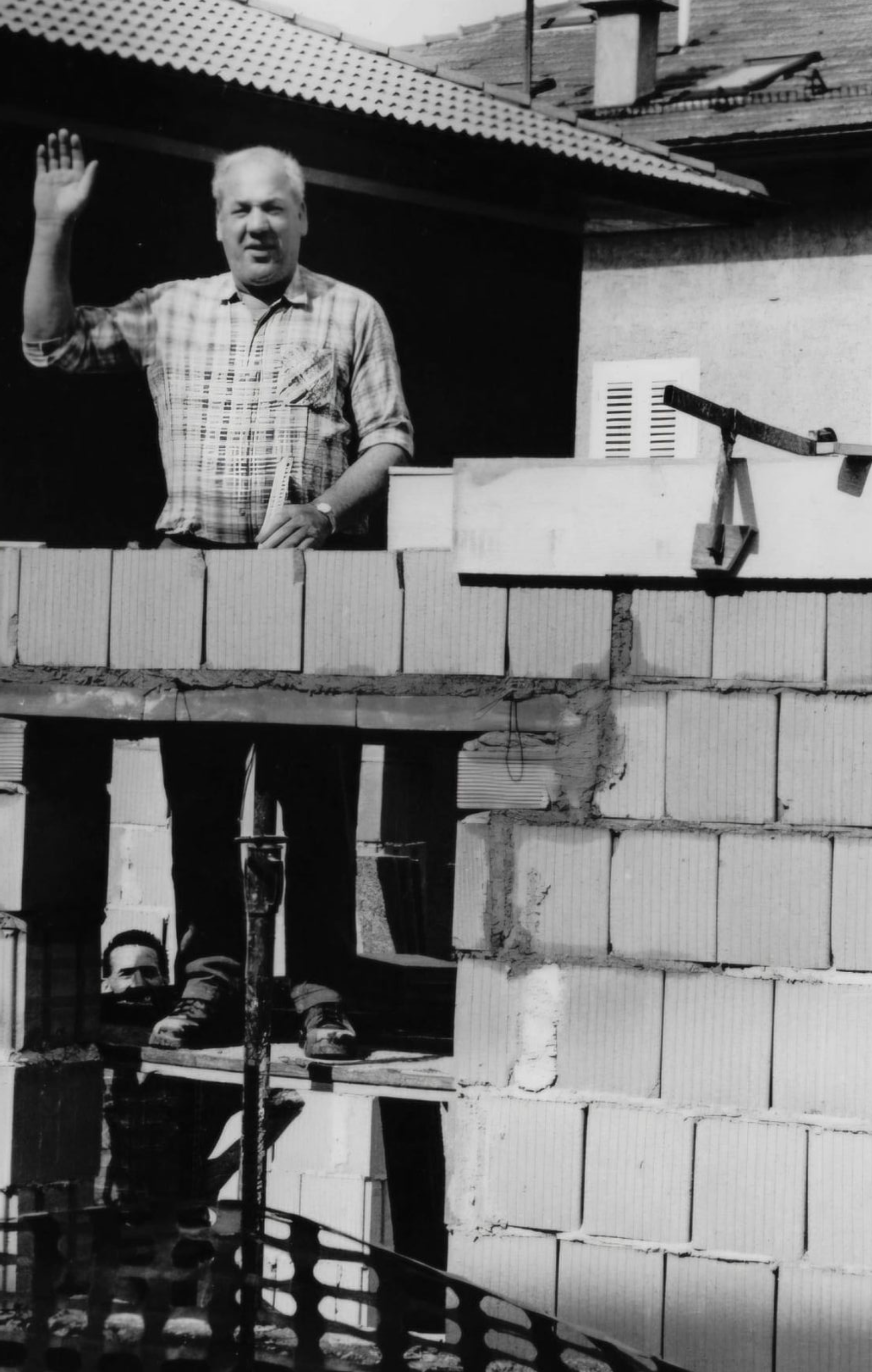 Un uomo saluta da una finestra sopra una struttura in mattoni parzialmente costruita, altre due persone più in basso.