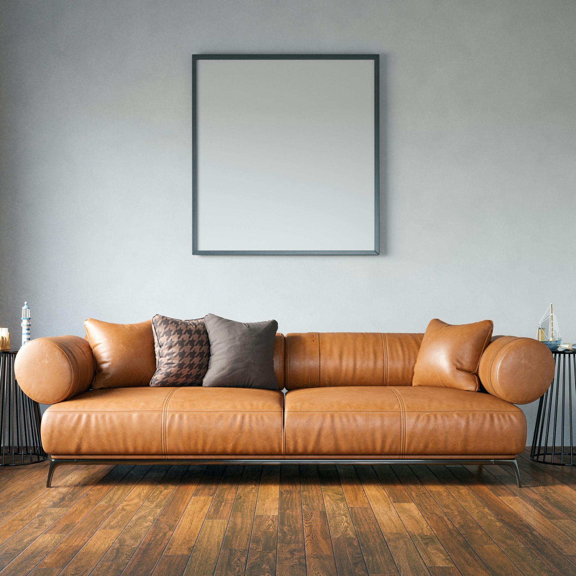Tan leather sofa with pillows beneath a framed blank picture on a gray wall.