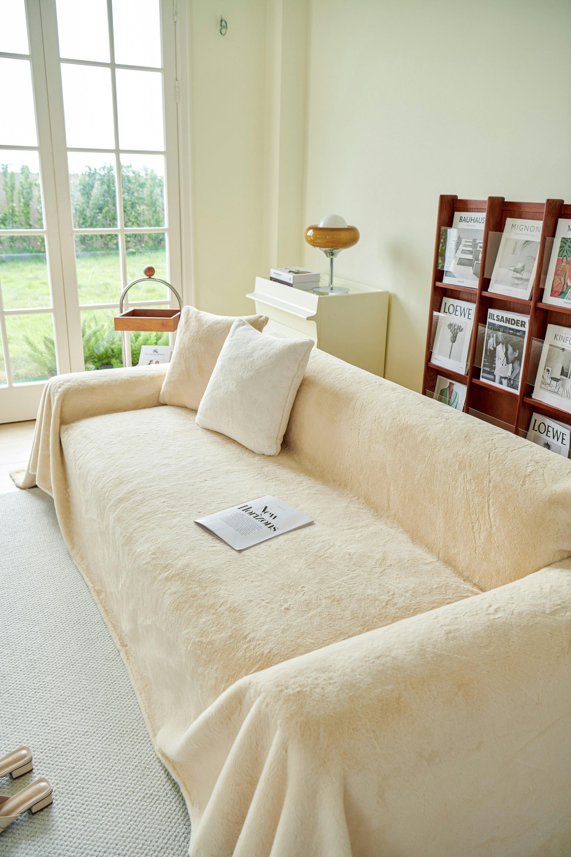 Beige sofa with a fluffy cover and pillows, next to a window and a bookshelf.