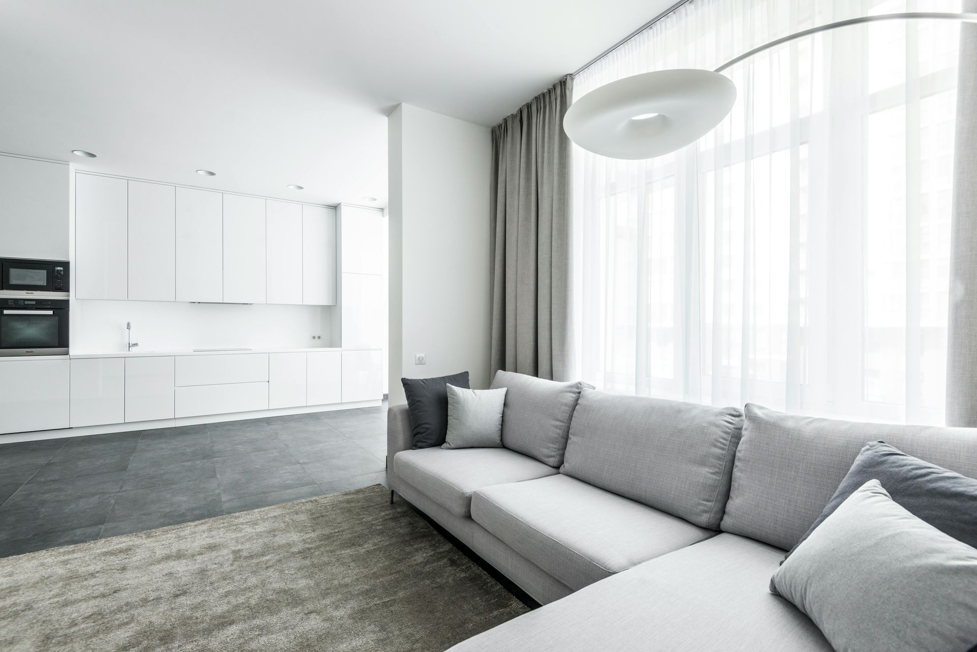 Modern living room with gray sectional sofa, white kitchen, and large window.