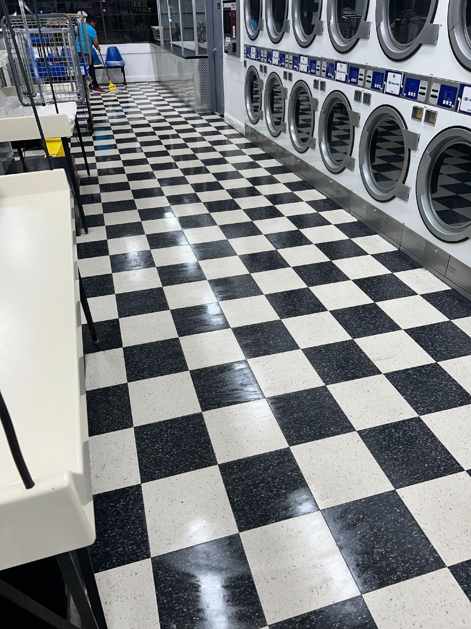 Black and white checkered floor of a laundromat with rows of washers and a person in the background.