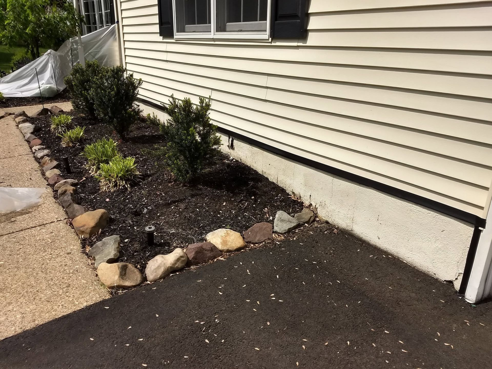 Front of a house with a mulched flower bed, shrubs, and a paved driveway beside beige siding.