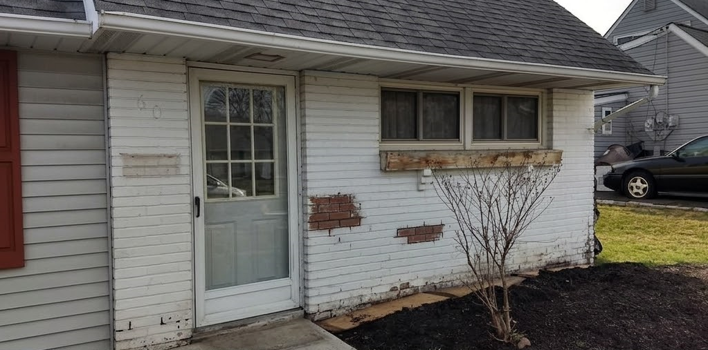 Damaged stucco foundation under tan siding beside a brick wall and paved patio