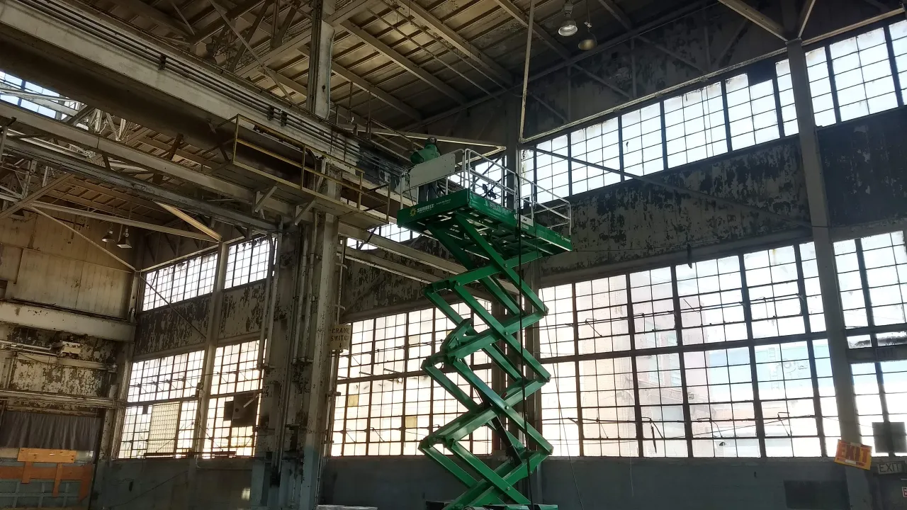 Green scissor lift in a large industrial warehouse with tall windows and exposed beams