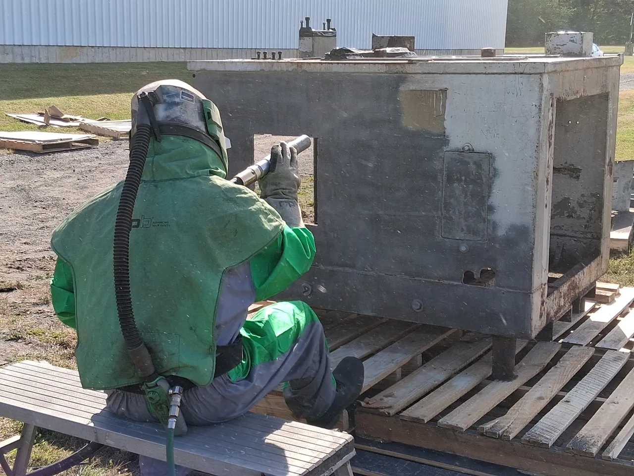 Worker in green protective gear sprays a large metal container on a wooden pallet outdoors.