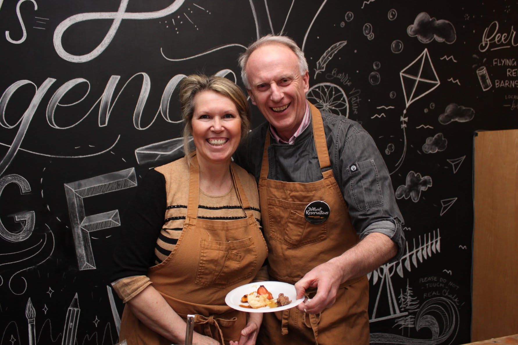 Smiling couple in brown aprons holding a plate of food in front of a chalkboard wall