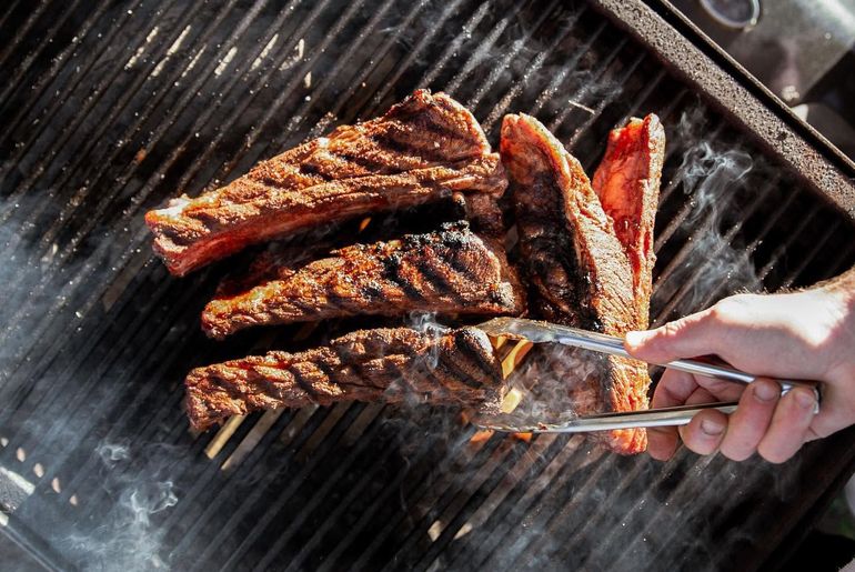 Ribs sizzling on a grill as a hand turns them with metal tongs, smoke rising.