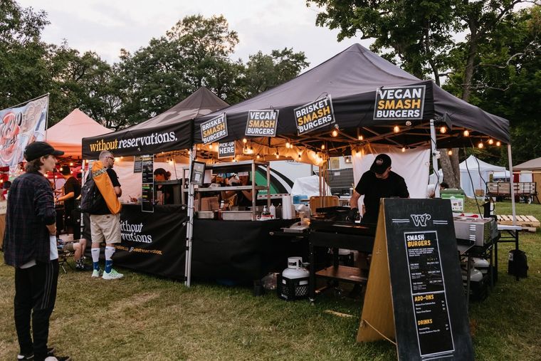 Outdoor vegan food stall with black canopies, menu boards, and customers at a grassy market.