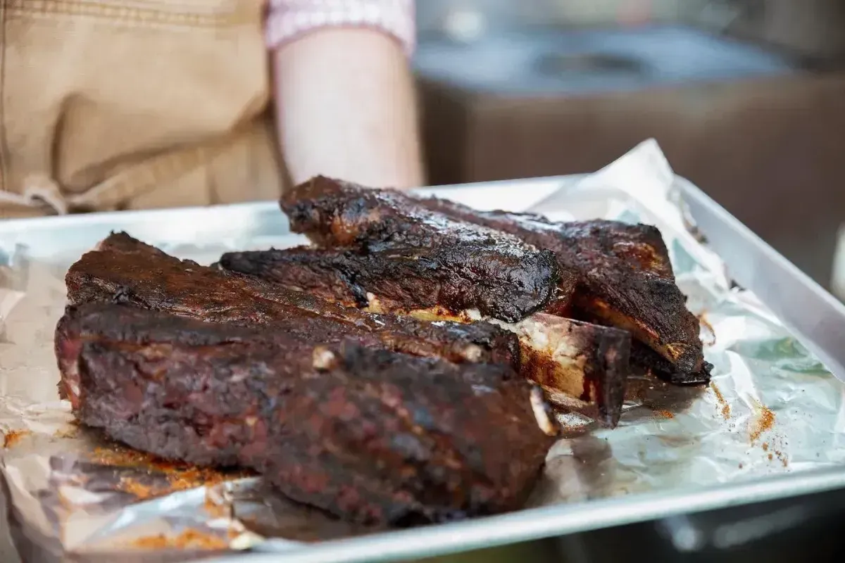 Tray of charred grilled meat on foil, with a person holding it in the background