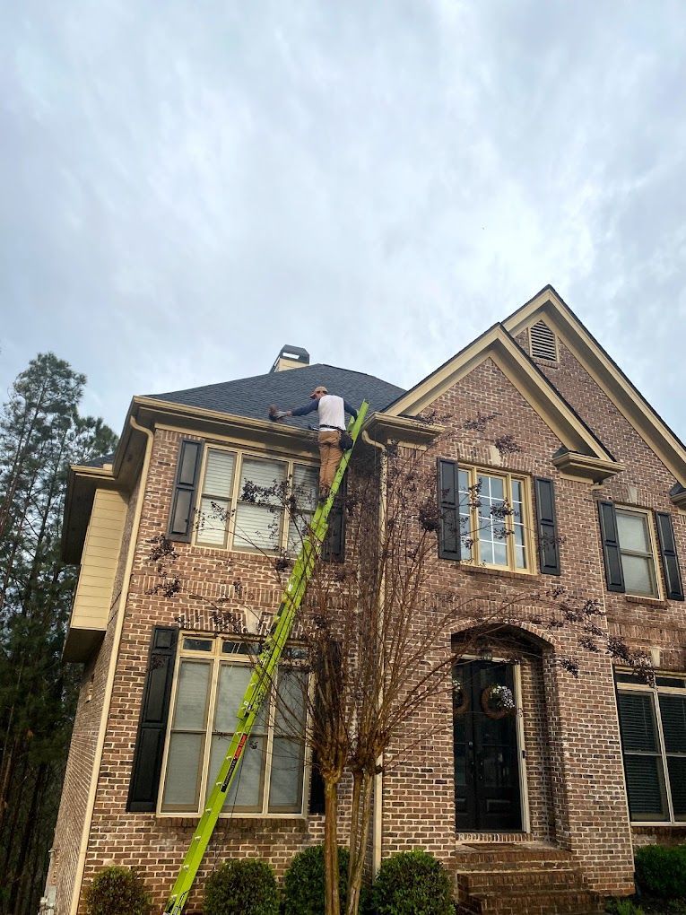 Person on a tall ladder cleaning gutters on a brick house under a cloudy sky.