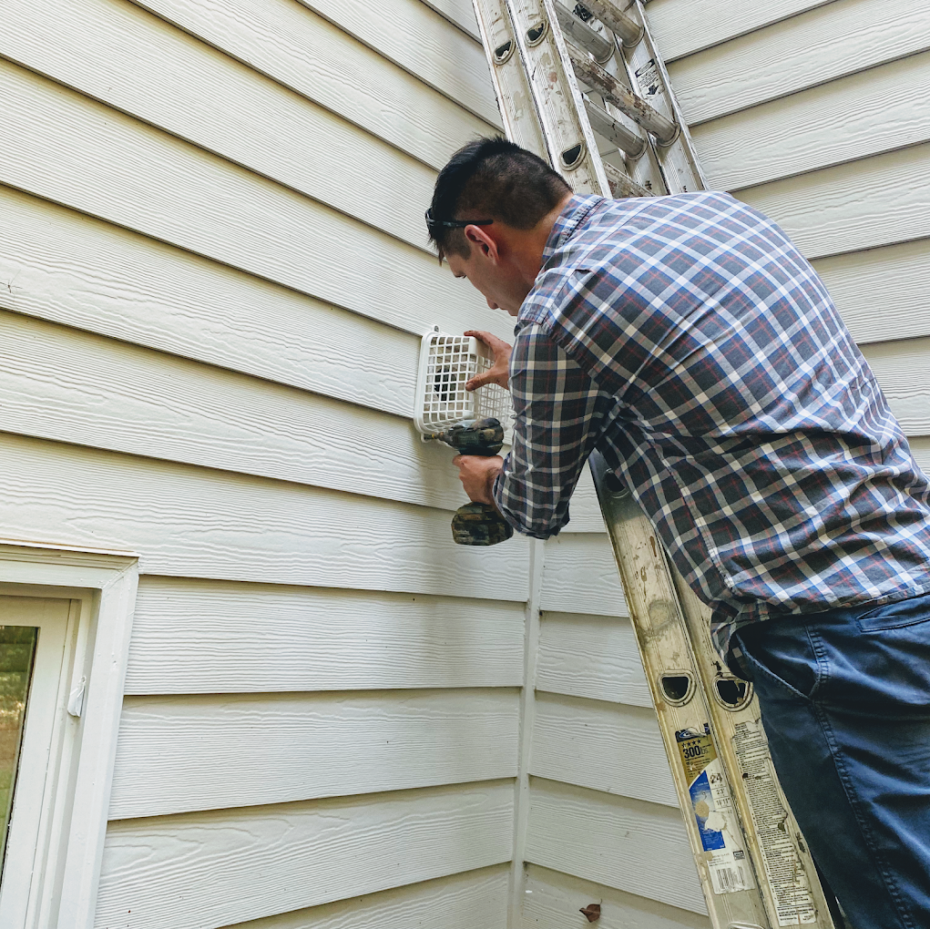 Man on a ladder, installing a vent on the exterior of a house.