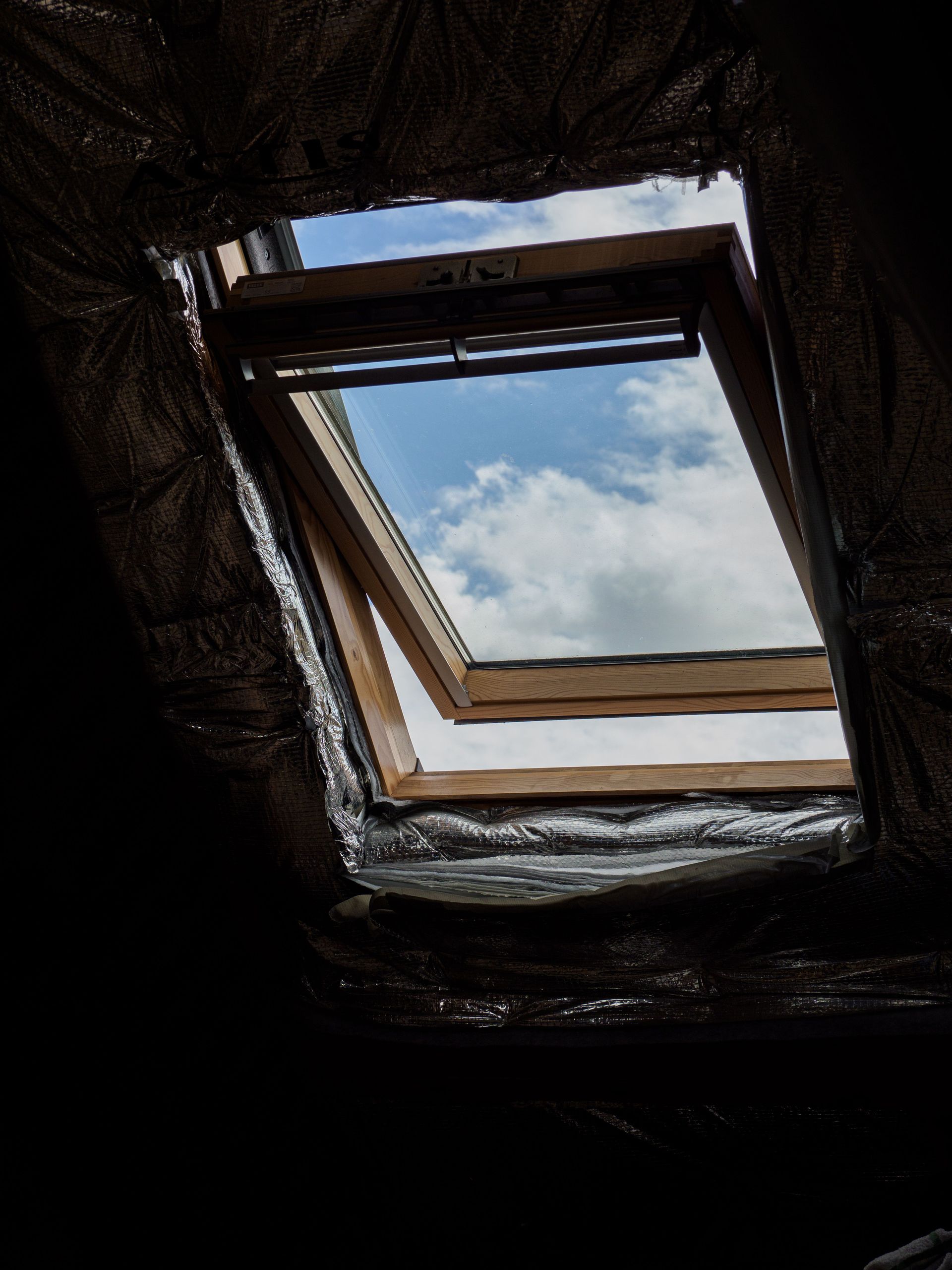 View through a square attic skylight opening to a cloudy sky, surrounded by dark insulation
