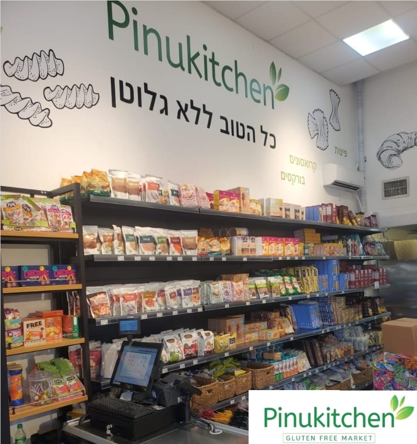 Pinukitchen grocery aisle with shelves of packaged foods and Hebrew signage, checkout counter in front
