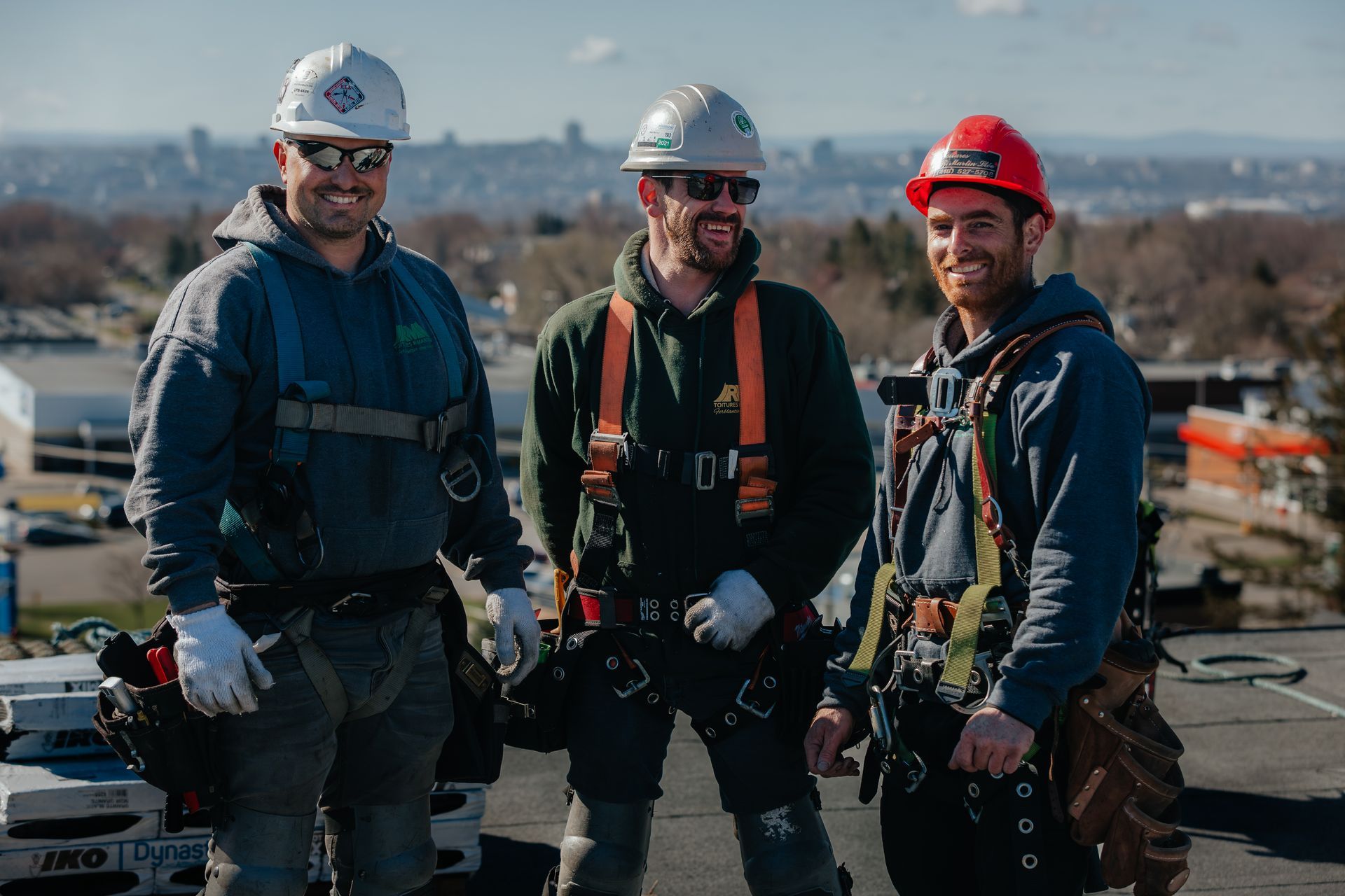 Trois ouvriers du bâtiment en tenue de sécurité, debout sur un toit, sourient à l'objectif.
