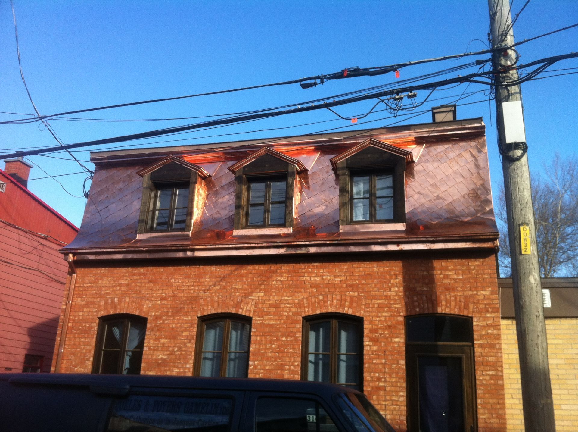 Maison de ville en briques avec toit rouge et lucarnes derrière une voiture garée dans une rue ensoleillée