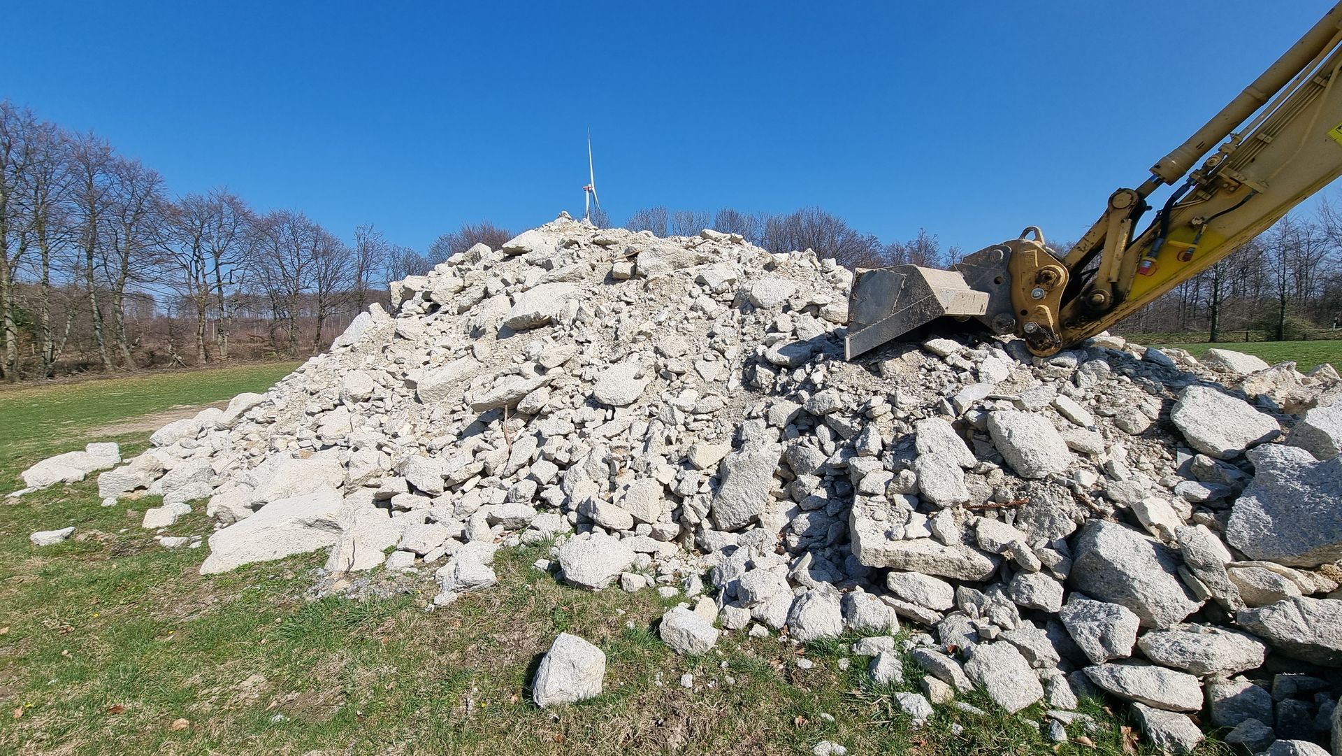 Excavator arm at a large pile of broken white rocks on a grassy hillside under a clear blue sky