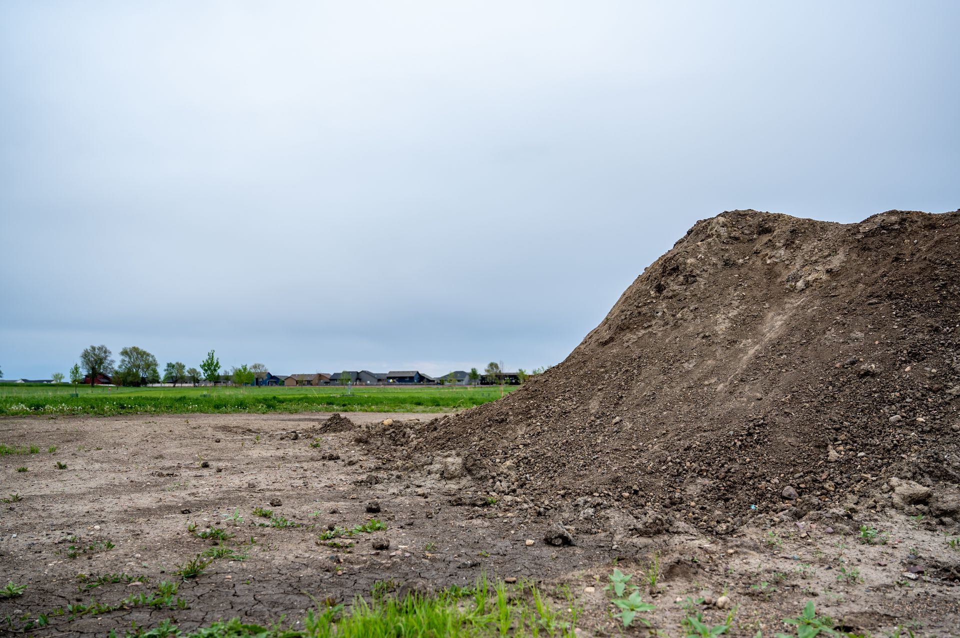 Large dirt mound in a bare field under a cloudy sky, with green farmland in the distance