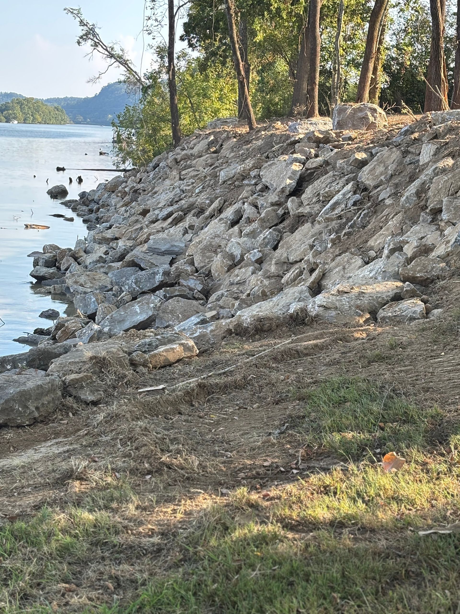 Rocky shoreline beside a calm lake, with trees and grass along the bank. Rocky shoreline beside a calm lake, with trees and grass along the bank.