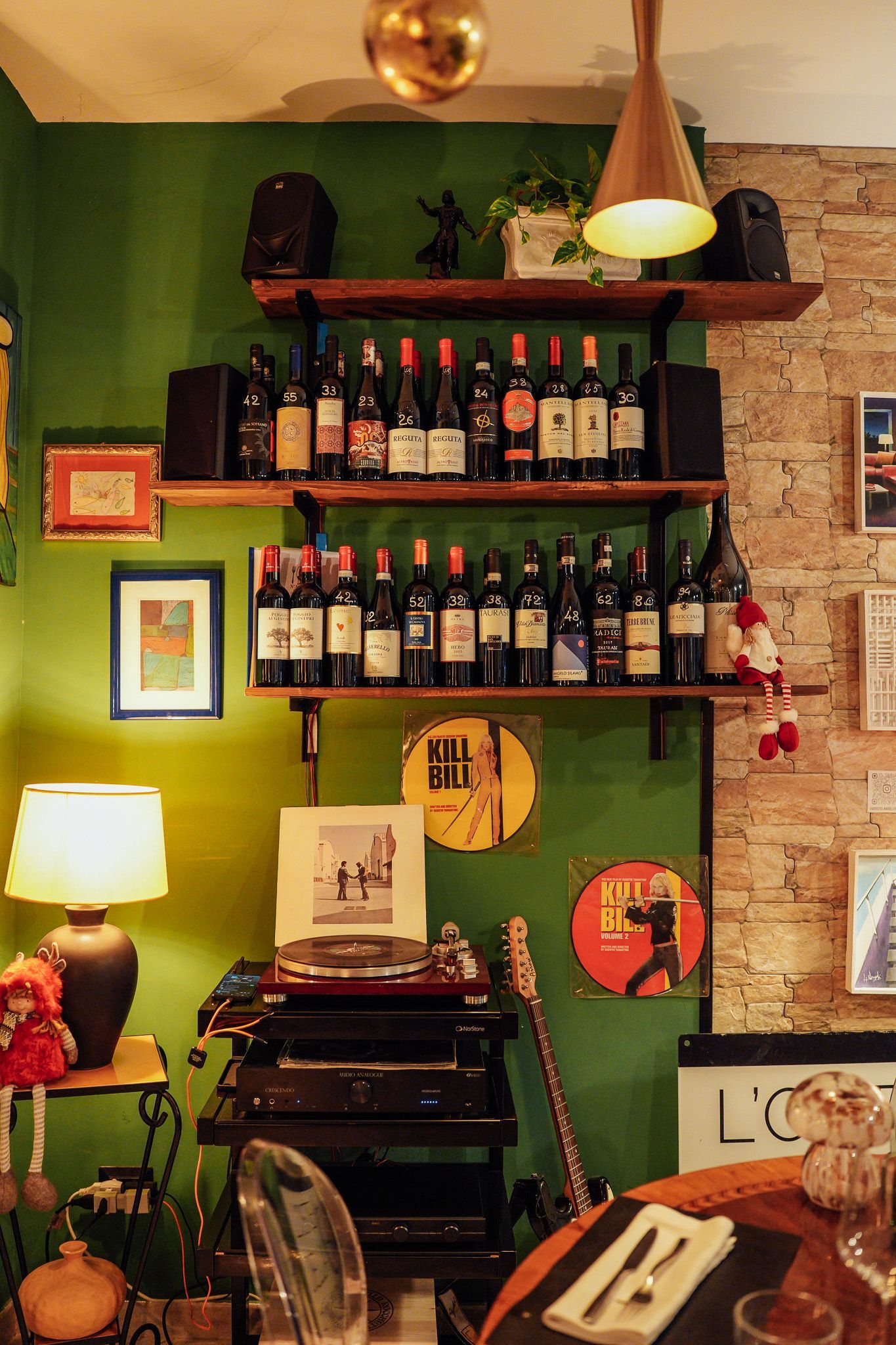 Interno di un ristorante con pareti verdi, dettagli in mattoni, scaffali pieni di vino e apparecchiature audio.