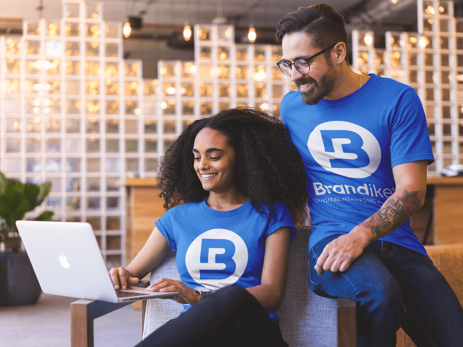 Two people in blue branded shirts smiling at a laptop in a warm, modern office