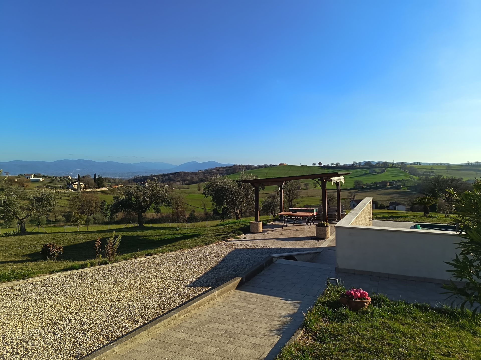 Un patio di ghiaia con pergolato in legno si affaccia su un paesaggio pittoresco e soleggiato 
