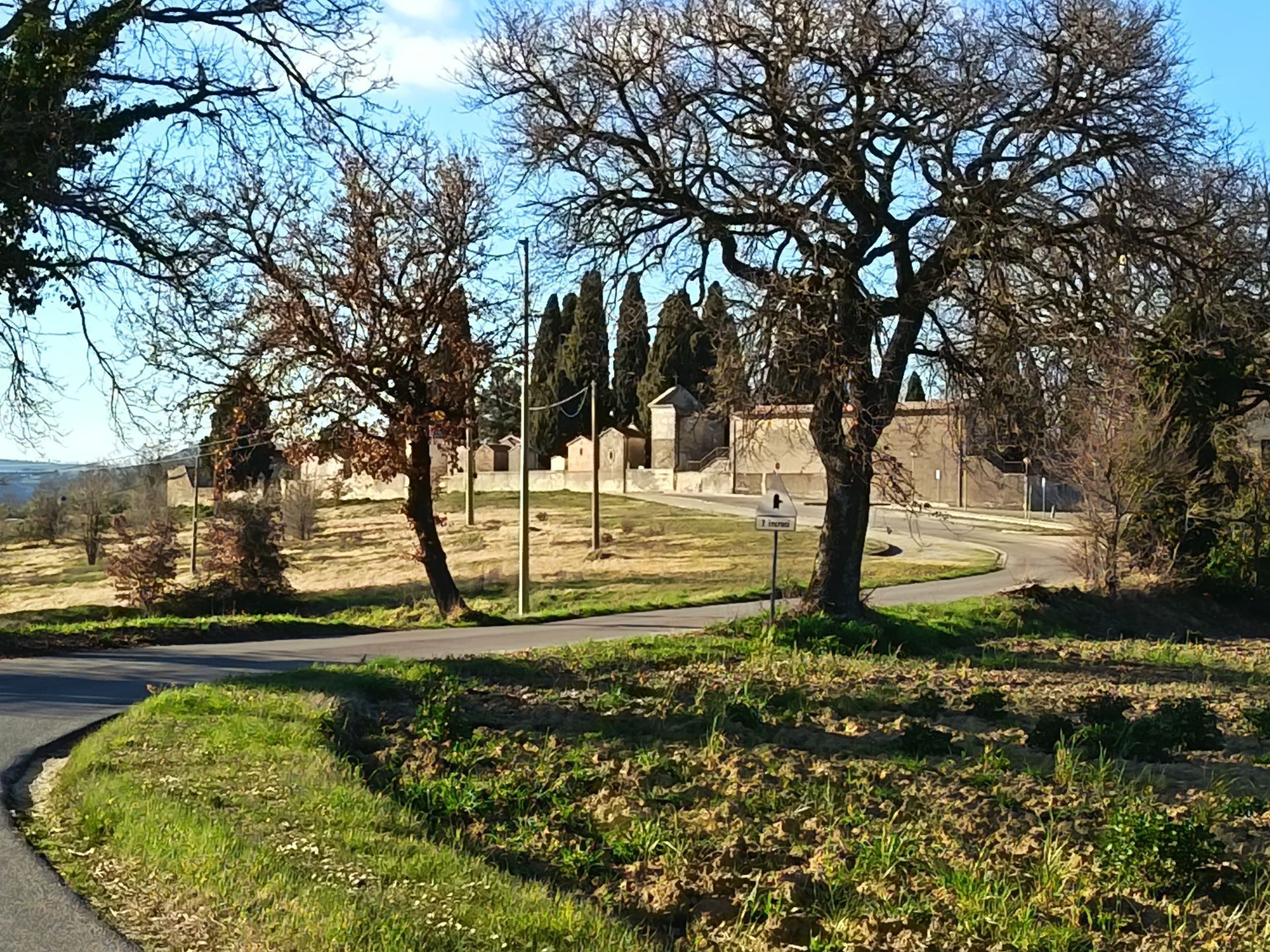 Una strada di campagna conduce a un edificio con muri in pietra, circondato da alberi spogli sotto un cielo limpido.