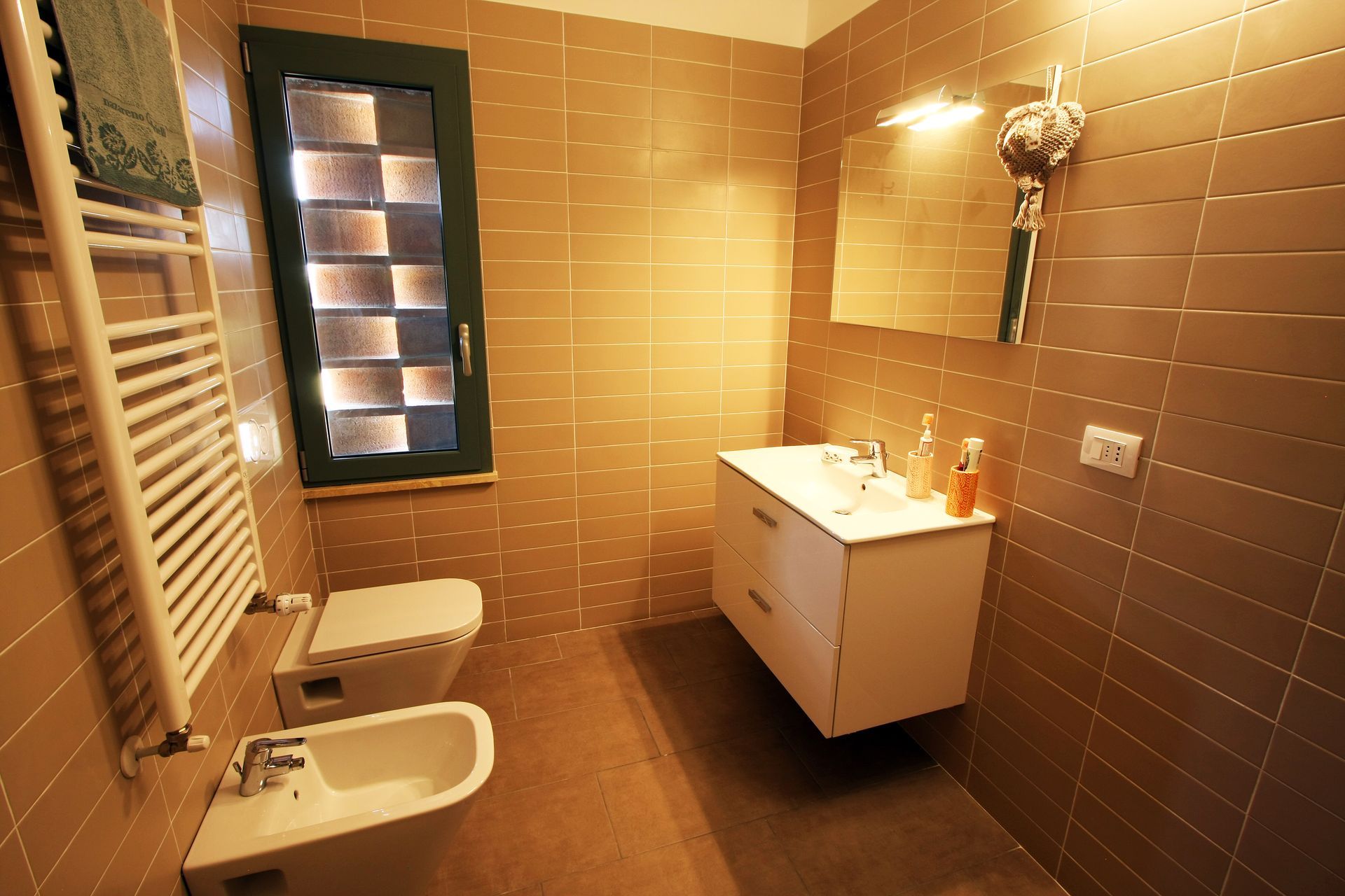 Un bagno moderno piastrellato con mobile lavabo bianco dotato di specchio, bidet, WC sospeso e scaldasalviette bianco.