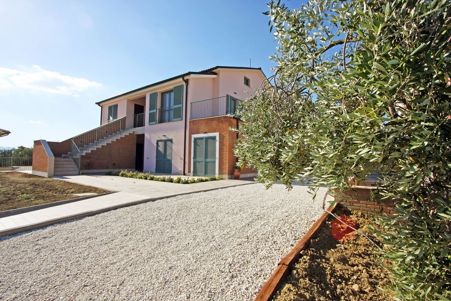 Una casa a due piani color rosa chiaro con dettagli in mattoni, una scala esterna e persiane verdi, accanto a un ulivo.