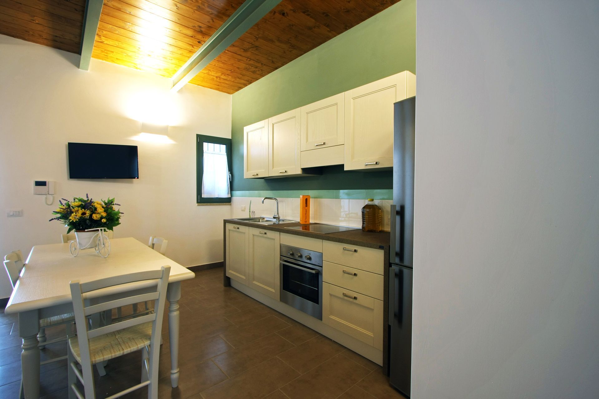 Una cucina con soffitto in legno, mobili chiari, forno in acciaio inox, tavolo da pranzo e televisore a parete.