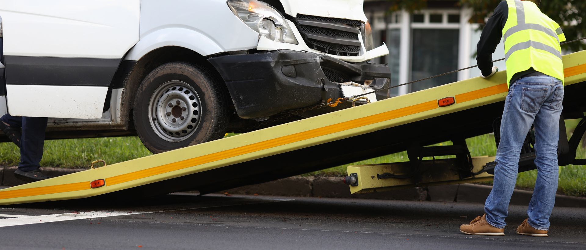 Damaged white van being winched onto a tow truck ramp by a worker in a safety vest.
