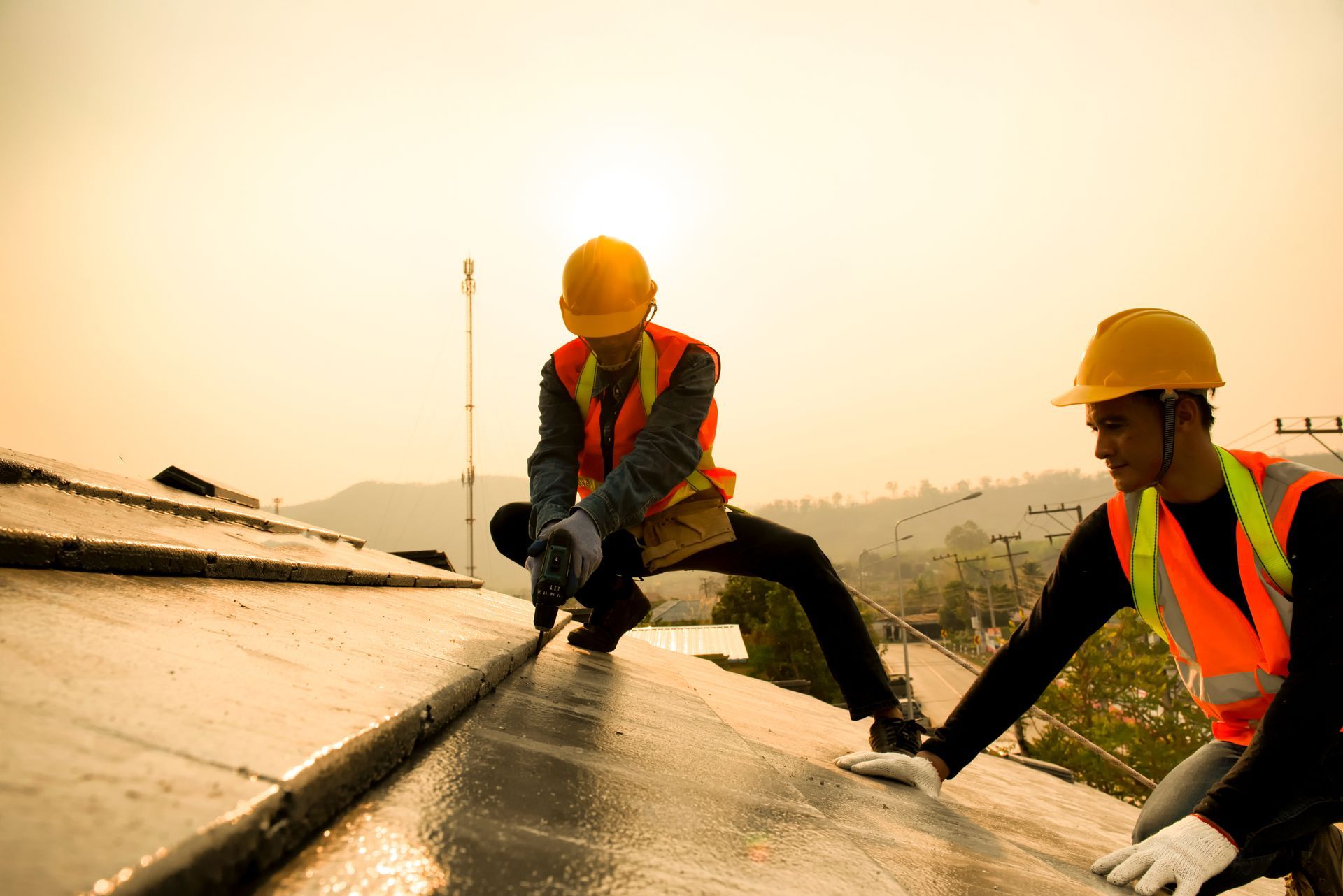 Two roofers in safety gear applying sealant to a roof at sunset.