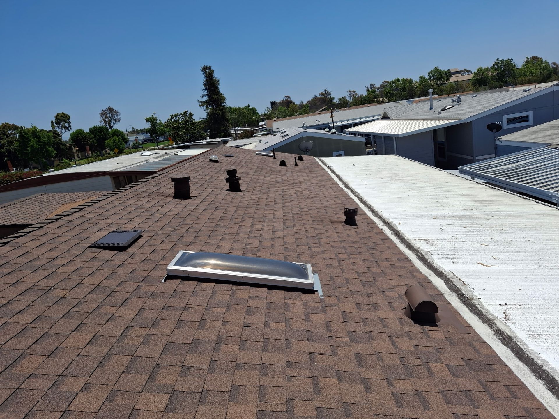 View across a rooftop with brown shingles, skylights, vents, and white flat-roof sections under a clear sky.