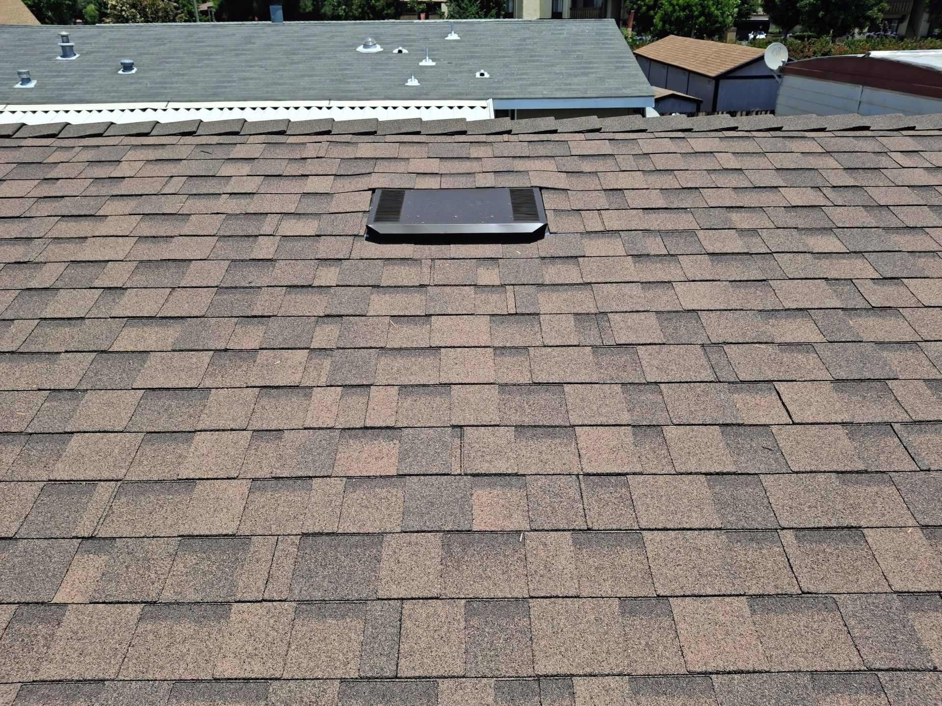 Rooftop with gray-brown shingles and a central skylight, with neighboring houses in the background