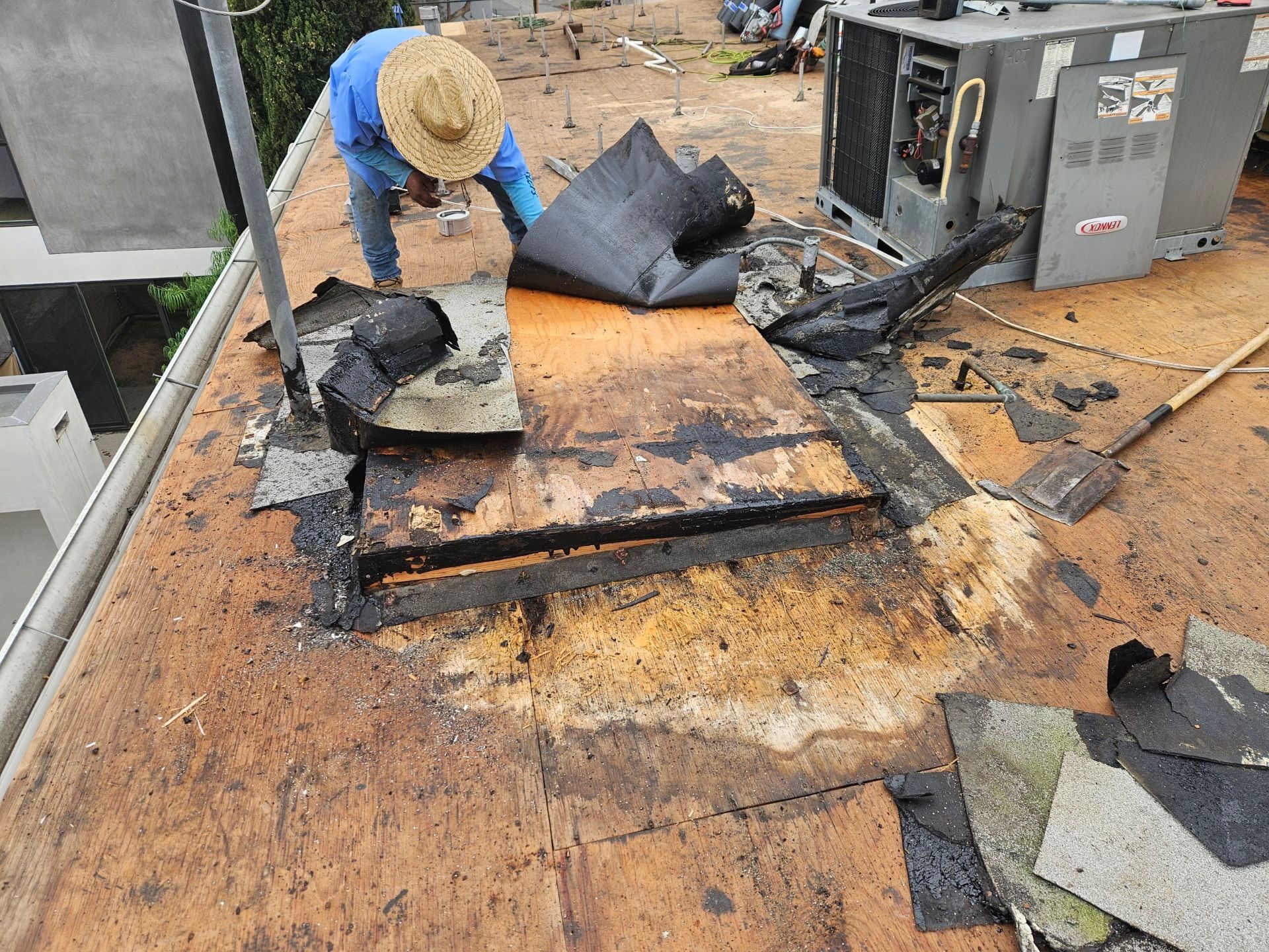 Worker removing torn roofing on a flat roof beside HVAC equipment.