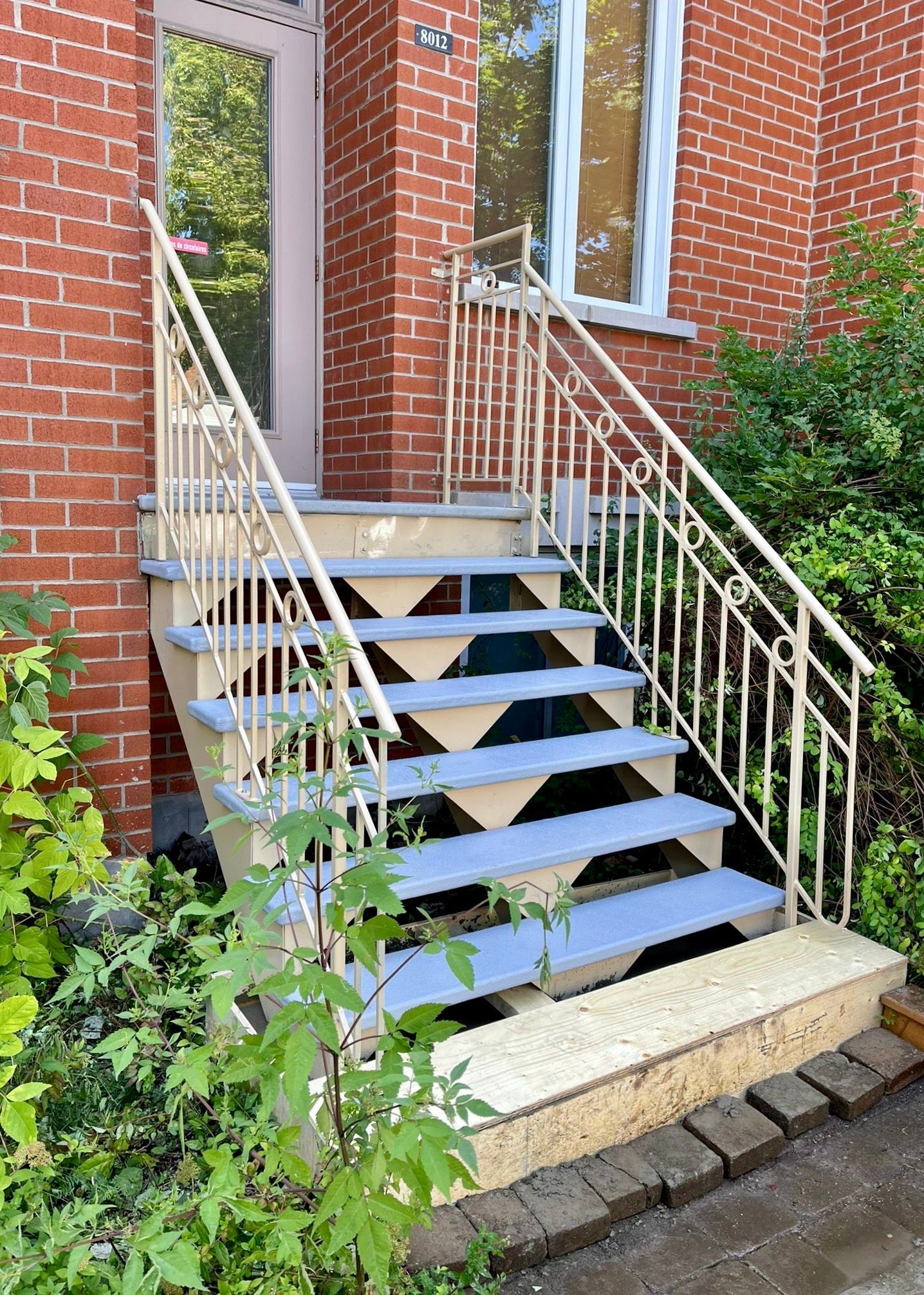Escalier extérieur avec rampe beige, menant à l'entrée d'un bâtiment en briques. Les marches sont peintes en bleu clair.