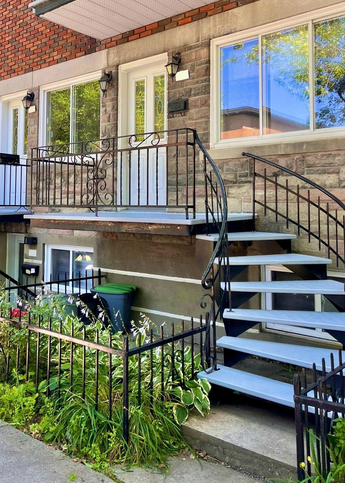 L'extérieur de cette maison en pierre présente une porte d'entrée blanche surélevée, une balustrade de balcon en métal et un escalier descendant vers une pelouse.