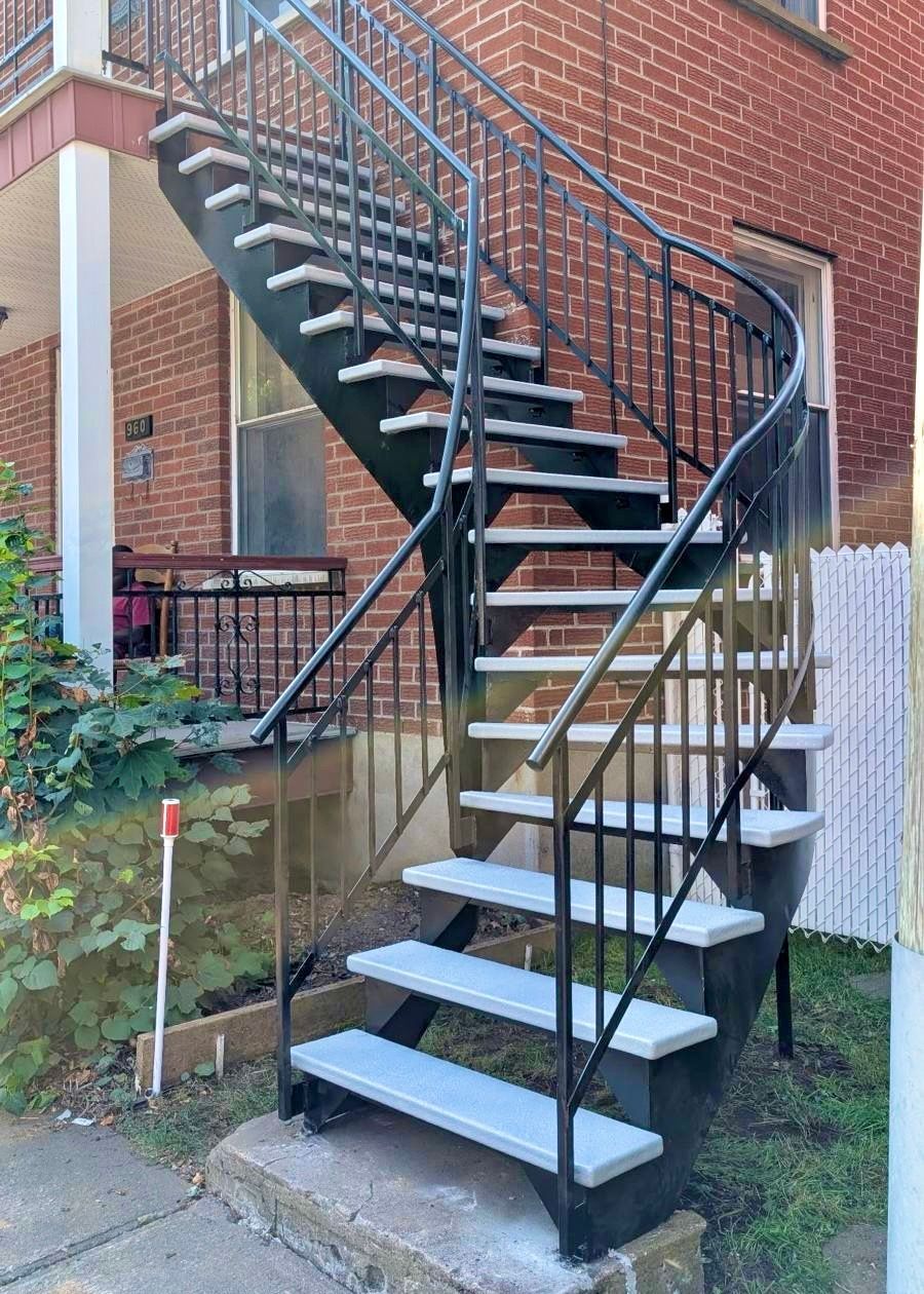 Escalier extérieur en métal avec rampes noires et marches grises, fixé à un bâtiment en briques rouges.