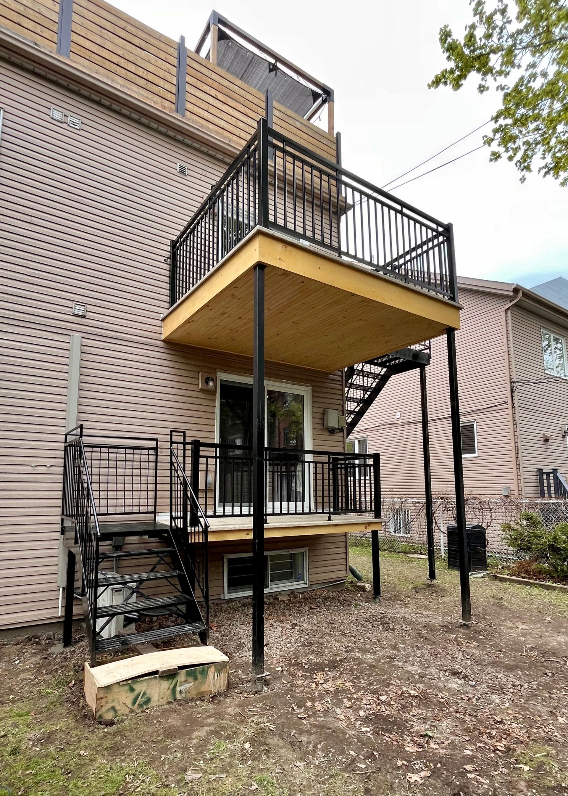 Terrasse métallique noire à deux étages avec escalier, rattachée à un bâtiment en briques. La terrasse au rez-de-chaussée se trouve au-dessus d'un escalier menant à la cour.