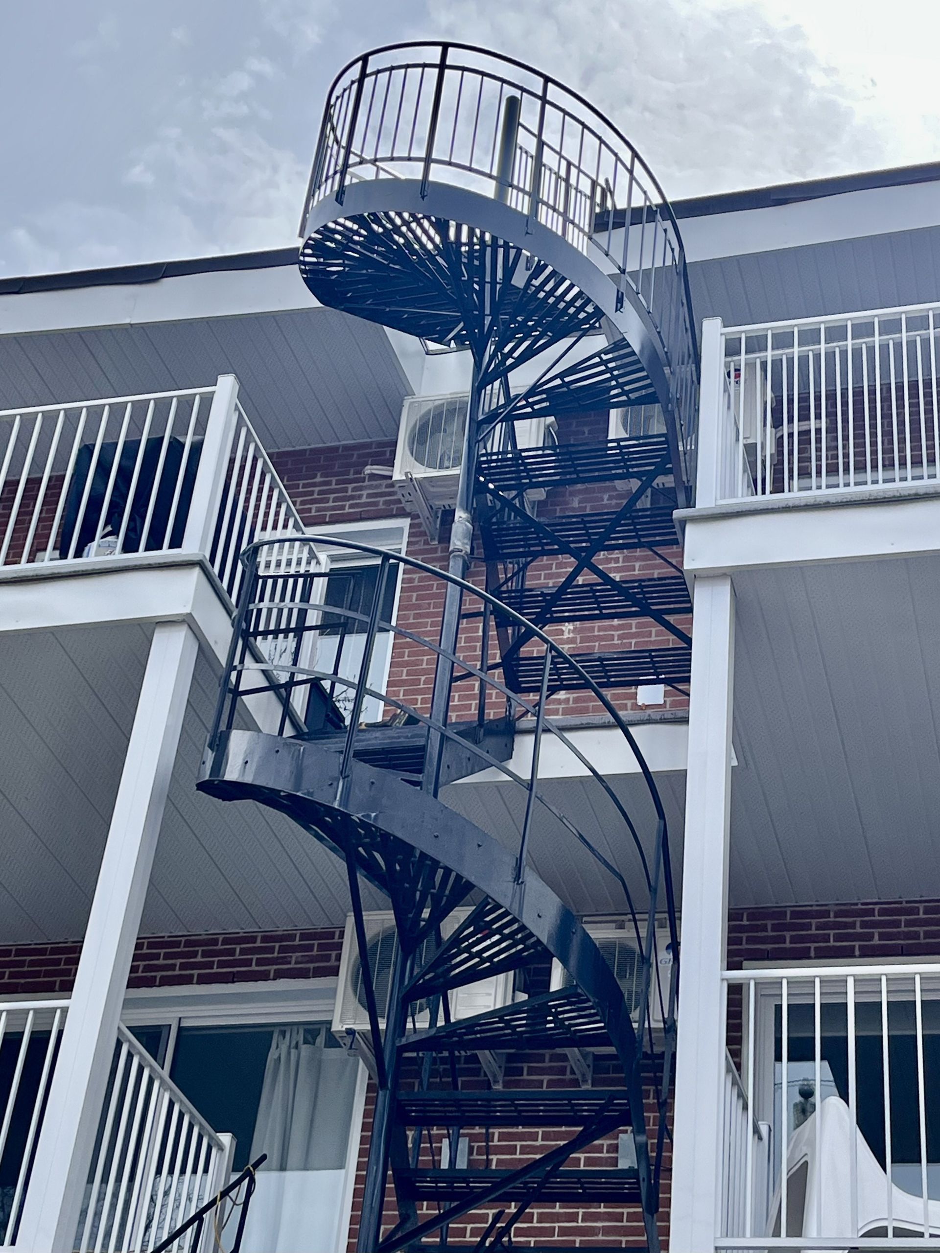 Un escalier en colimaçon noir à l'extérieur d'un bâtiment en briques, avec des balcons et une unité de climatisation visibles.