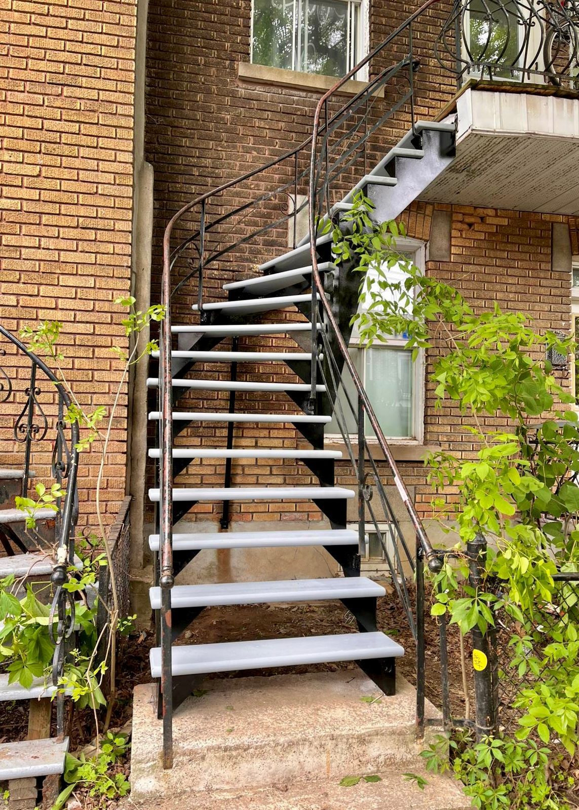 Escalier extérieur en métal adossé à un bâtiment en briques, avec des vignes et une fenêtre visibles.